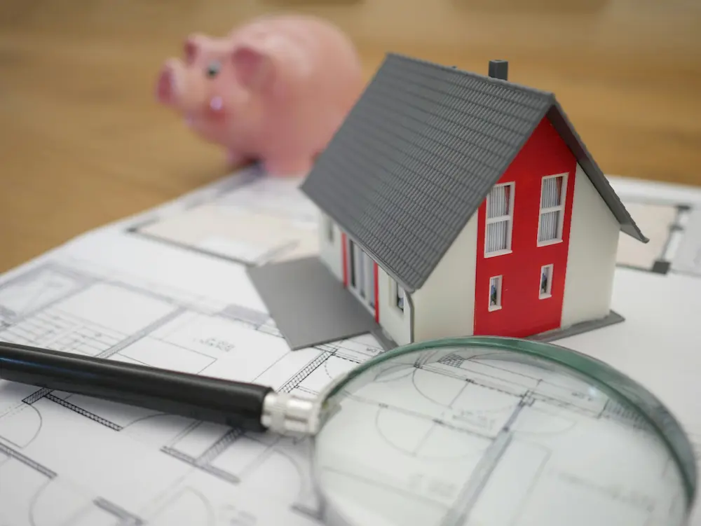 Miniature house and magnifying glass on top of blue prints with piggy bank in the background